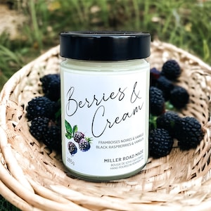 May include: A white candle jar with a black lid, sitting in a wicker basket filled with blackberries. The label reads "Berries & Cream", "Framboises Noires & Vanille", "Black Raspberries & Vanilla", "Miller Road Made", "Bougie de Soja | Soy Candle", "Hand Poured in Renfrew, ON", and "285g".