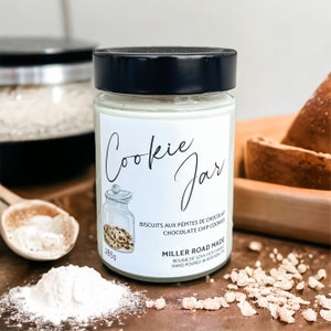 May include: A glass jar with a black lid, labelled "Cookie Jar." The label reads "BISCUITS AUX PÉPITES DE CHOCOLAT CHOCOLATE CHIP COOKIES" and "MILLER ROAD MADE." The jar contains a white candle and is surrounded by cookie ingredients, including flour.