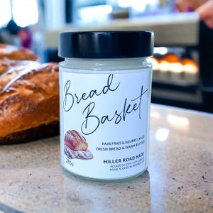 May include: A white candle jar with a black lid. The label reads "Bread Basket" in cursive script. The candle is a soy candle and is hand-poured. The label also says "Pain frais & beurre chaud" and "Fresh bread & warm butter".