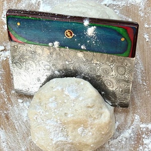 Puede incluir: Primer plano de una bola de masa espolvoreada con harina, siendo cortada con un cortador de masa de acero inoxidable con un mango de madera de colores.