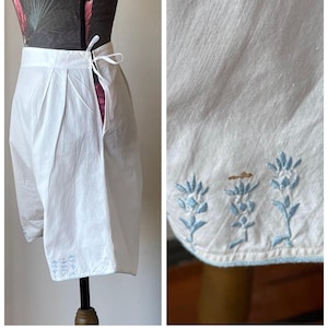 Könnte beinhalten: Weiße Baumwollshorts mit Falten und Kordelzug in der Taille. Die Shorts haben hellblaue Blumenstickereien am Saum. Die Shorts werden auf einer Schaufensterpuppe präsentiert.