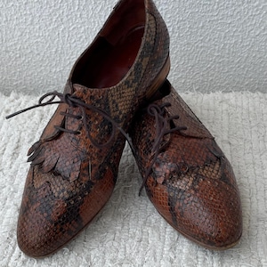 Könnte beinhalten: Ein Paar braune Lederschuhe mit Schlangenmuster. Die Schuhe haben Schnürsenkel und dekorative Fransen an der Spitze. Die Schuhe sind Größe 42, etwa 23 cm.