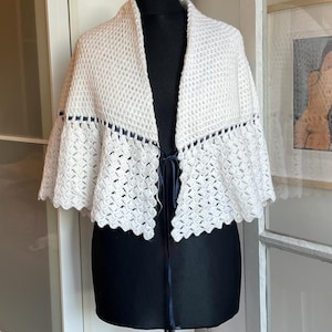 May include: A white, open-front crochet shawl with a scalloped edge and a navy blue ribbon tie. The shawl is displayed on a black mannequin, showcasing its textured, open-weave design. The garment appears handmade.