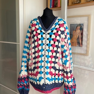 Peut inclure: Un pull à col en V et manches longues réalisé avec un motif au crochet de type granny square multicolore. Le vêtement présente un design répétitif de carrés en laine bleu marine foncé, turquoise vif, magenta éclatant et blanc cassé.