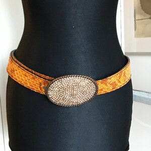 Puede incluir: Un cinturón marrón y beige con estampado de piel de serpiente. El cinturón presenta una hebilla ovalada grande adornada con pequeños pedrería brillantes. La hebilla está enmarcada por un diseño de cuerda trenzada.