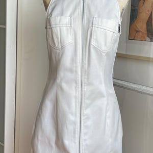 May include: White halter-neck dress with a full-length zipper and two front pockets. The dress is displayed on a mannequin. The dress has a fitted silhouette and a small logo patch near the hem. The dress is made of a smooth, possibly leather-like material.