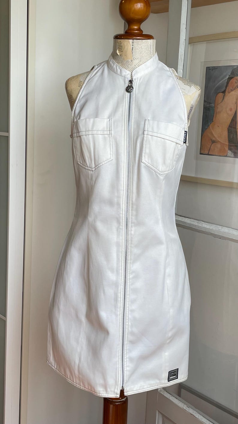 May include: White halter-neck dress with a full-length zipper and two front pockets. The dress is displayed on a mannequin. The dress has a fitted silhouette and a small logo patch near the hem. The dress is made of a smooth, possibly leather-like material.