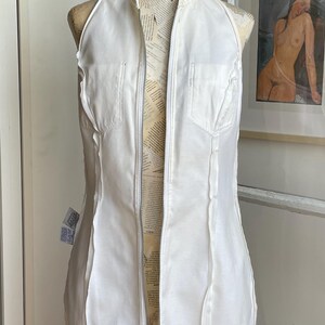 May include: A white halter-style dress with a full-length zipper down the front, displayed on a dress form. The dress has a fitted silhouette and features vertical seams and small pockets. The dress form is covered in newsprint.