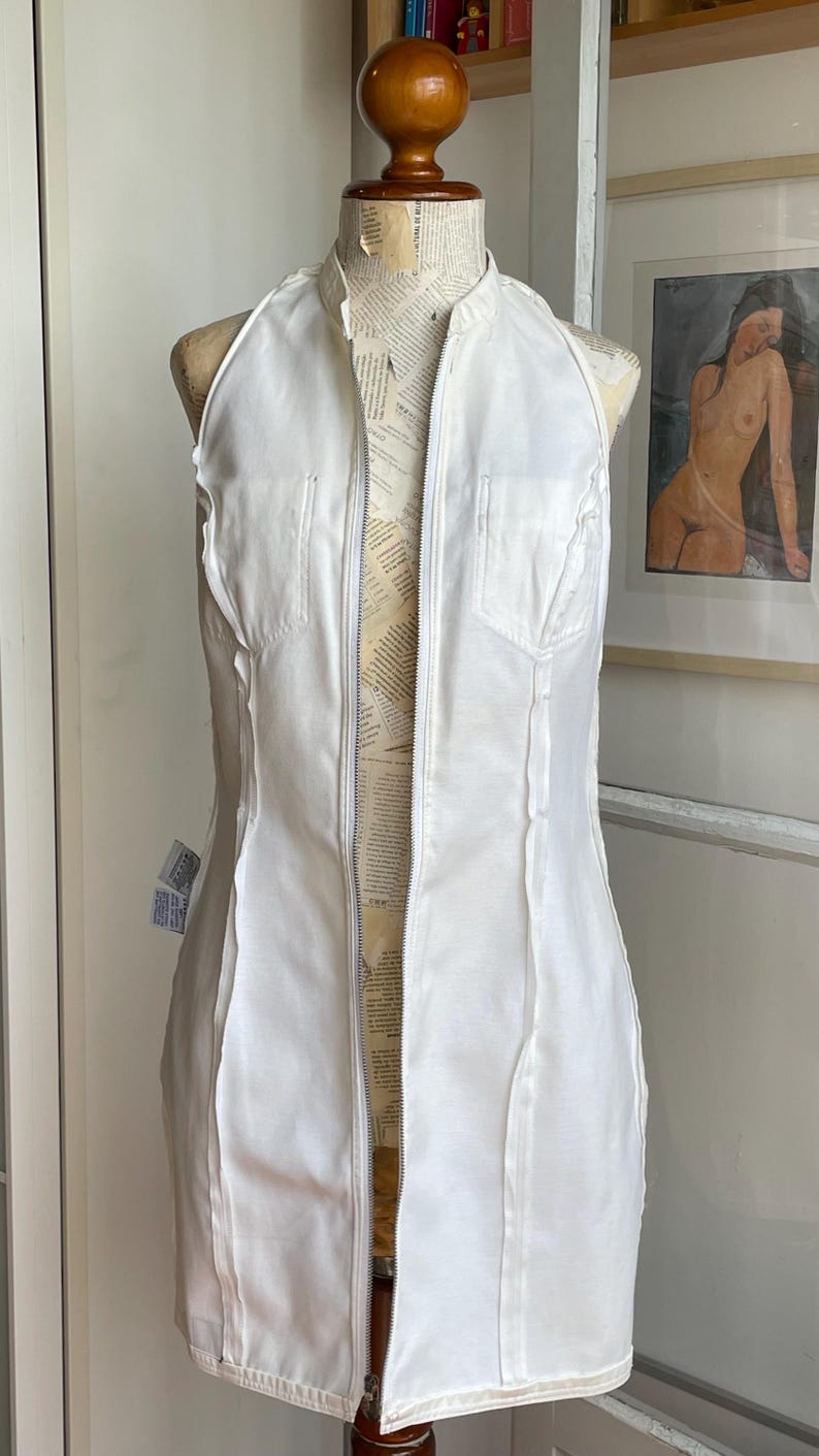 May include: A white halter-style dress with a full-length zipper down the front, displayed on a dress form. The dress has a fitted silhouette and features vertical seams and small pockets. The dress form is covered in newsprint.