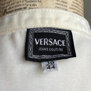 May include: Close-up of a white garment with a black Versace Jeans Couture label. The label features the brand name and a size tag with the numbers 30 and 44.
