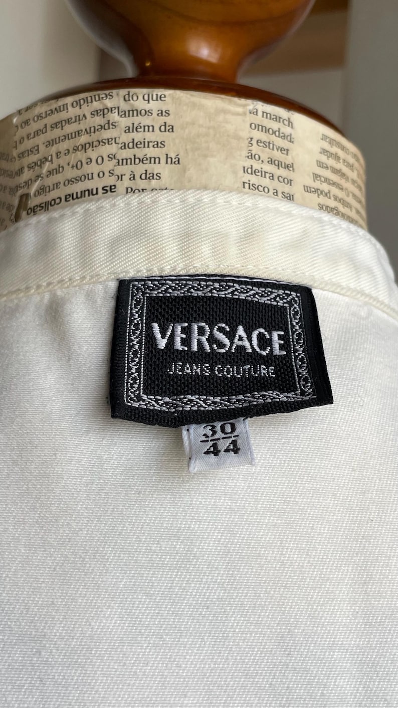 May include: Close-up of a white garment with a black Versace Jeans Couture label. The label features the brand name and a size tag with the numbers 30 and 44.