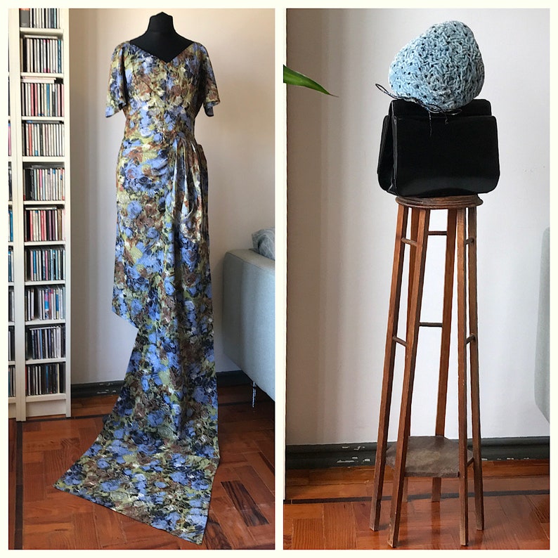 May include: A long, flowing dress with a blue and green floral pattern. The dress is on a mannequin and is displayed in a room with a bookshelf and a couch.