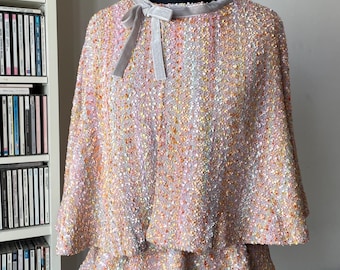 Puede incluir: Un top tipo capa de tweed multicolor con un dobladillo con volantes y un lazo de cinta de terciopelo gris en el cuello. El top se exhibe en un maniquí negro y tiene una apariencia texturizada.