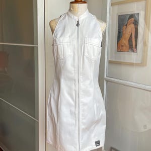 May include: White halter-neck dress with a zip-up front and two chest pockets. The dress is displayed on a wooden mannequin. The dress has a small black and white tag near the hem. The dress is made of a smooth, possibly faux leather material.
