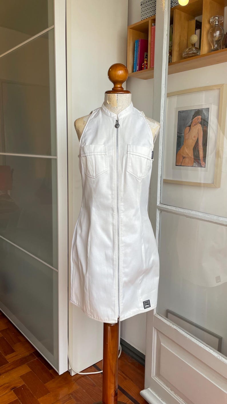 May include: White halter-neck dress with a zip-up front and two chest pockets. The dress is displayed on a wooden mannequin. The dress has a small black and white tag near the hem. The dress is made of a smooth, possibly faux leather material.