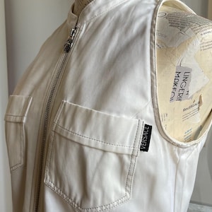 May include: Cream-colored sleeveless jacket with a high collar and a silver zipper. Features two front pockets and a small black Versace label. The jacket is displayed on a mannequin.