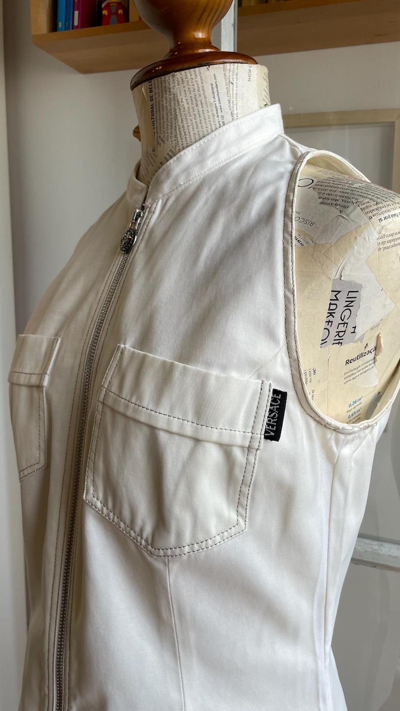 May include: Cream-colored sleeveless jacket with a high collar and a silver zipper. Features two front pockets and a small black Versace label. The jacket is displayed on a mannequin.