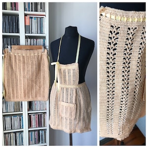 May include: A crocheted apron with a yellow ribbon trim and a pocket. The apron is beige and has a delicate, lacy pattern.