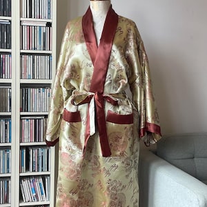 Puede incluir: Una bata de manga larga en tejido de satén dorado pálido con un patrón repetitivo de dragones rojos y dorados oscuros. La prenda incluye un cuello chal de color rojo oscuro sólido, puños a juego, dos bolsillos delanteros y un cinturón de lazo.
