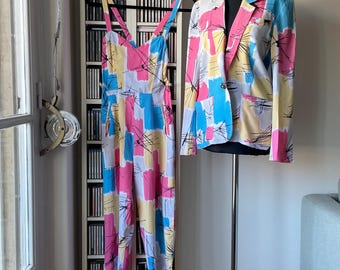May include: A colorful jumpsuit and blazer set. The jumpsuit has a V-neck and wide legs, with a pattern of pink, blue, yellow, and white rectangles. The blazer, on a mannequin, has a matching pattern. The clothing is displayed in a room with a CD rack.