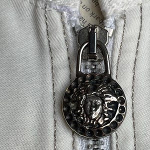May include: Close-up of a silver zipper pull with a Medusa head design on a white garment. The zipper is open, revealing the teeth and a portion of the fabric. The garment has silver stitching.