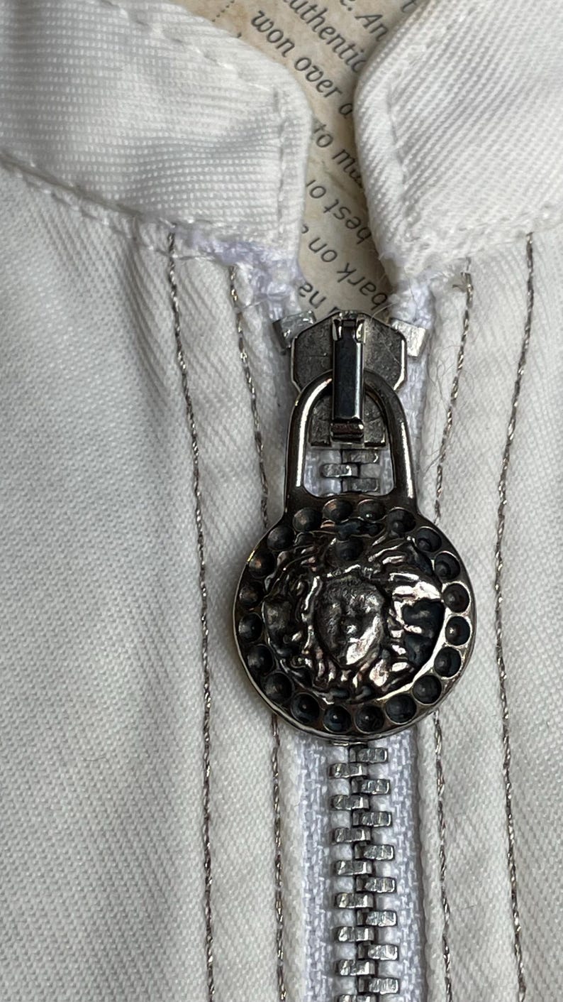 May include: Close-up of a silver zipper pull with a Medusa head design on a white garment. The zipper is open, revealing the teeth and a portion of the fabric. The garment has silver stitching.
