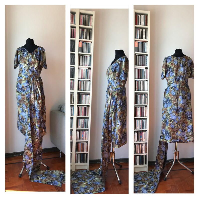 May include: A blue and brown floral print dress with a long train. The dress is on a mannequin and is displayed in a room with bookshelves.