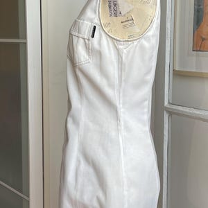 May include: A sleeveless white dress with a tailored fit, featuring a collared neckline and two front pockets. The dress is displayed on a mannequin, showcasing its clean lines and minimalist design. A small label is visible on the pocket.