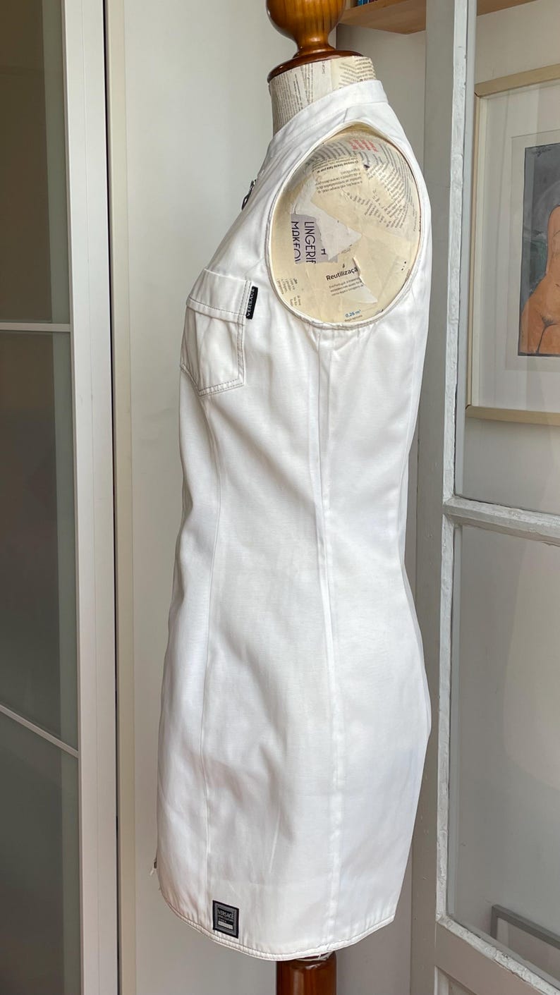 May include: A sleeveless white dress with a tailored fit, featuring a collared neckline and two front pockets. The dress is displayed on a mannequin, showcasing its clean lines and minimalist design. A small label is visible on the pocket.