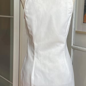 May include: A white, sleeveless dress with a mandarin collar, displayed on a mannequin. The dress has a fitted silhouette and a subtle sheen. The back of the dress is visible, showcasing its tailored design.