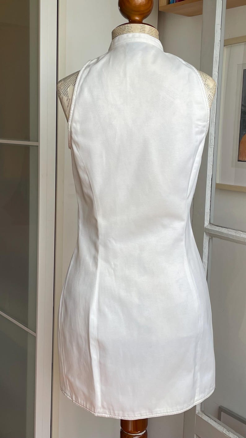 May include: A white, sleeveless dress with a mandarin collar, displayed on a mannequin. The dress has a fitted silhouette and a subtle sheen. The back of the dress is visible, showcasing its tailored design.