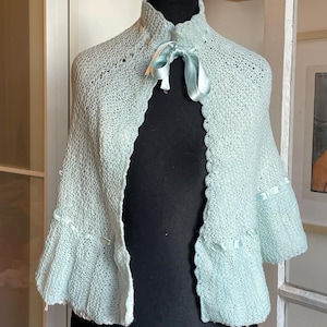 May include: A pale blue knitted cape with a ruffled hem and short sleeves. The cape has a high, rounded collar and is decorated with pale blue satin ribbon and a bow at the neckline. The garment is displayed on a black mannequin.