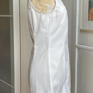 May include: A sleeveless white dress with a high collar and a front zipper. The dress features a single chest pocket and is displayed on a mannequin. The dress is made of a smooth, possibly cotton, fabric.