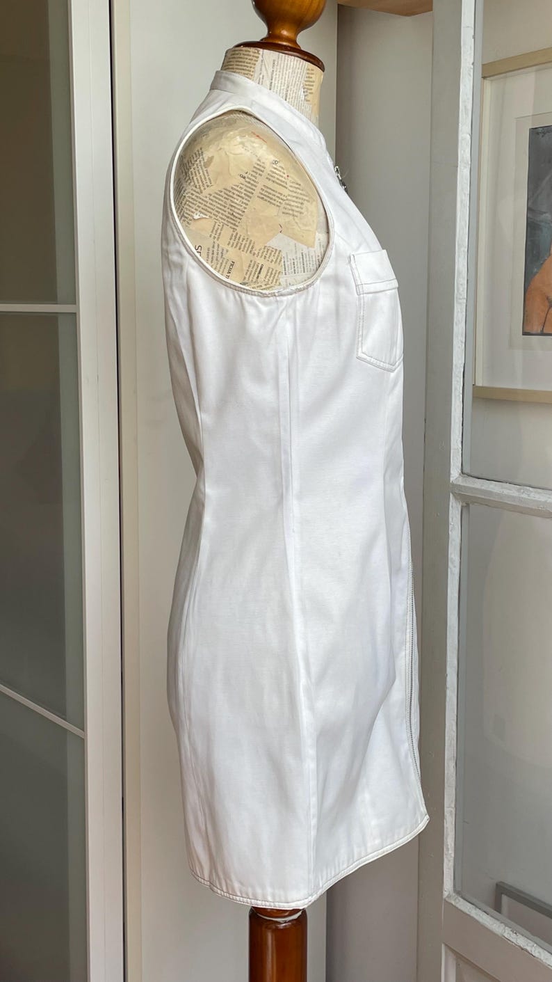 May include: A sleeveless white dress with a high collar and a front zipper. The dress features a single chest pocket and is displayed on a mannequin. The dress is made of a smooth, possibly cotton, fabric.