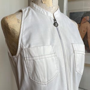 May include: A sleeveless white top with a mandarin collar and a full-length zipper. The top features two front pockets and is displayed on a mannequin. The stitching is visible in a contrasting color.