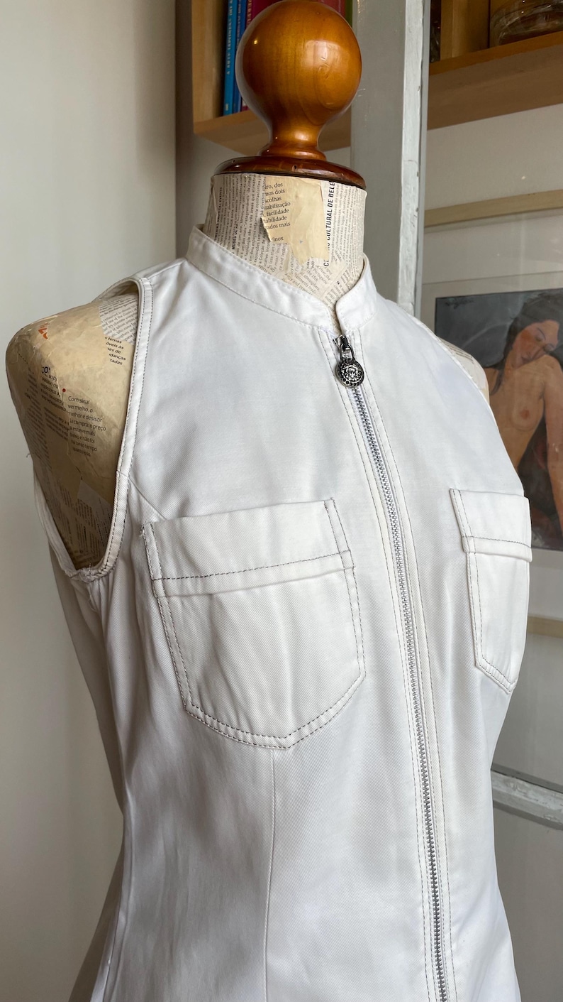 May include: A sleeveless white top with a mandarin collar and a full-length zipper. The top features two front pockets and is displayed on a mannequin. The stitching is visible in a contrasting color.