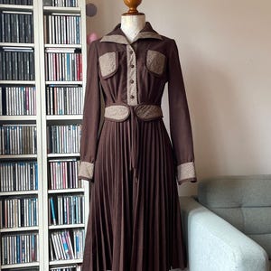 May include: A brown vintage dress with a pleated skirt and long sleeves. The dress features a collared neckline, button-down front, and pockets with a contrasting patterned trim. A matching belt cinches the waist. The dress is displayed on a wooden mannequin.