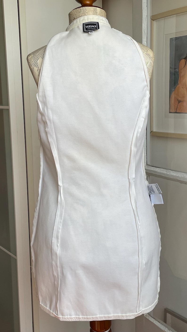 May include: A white sleeveless dress with a high neckline and vertical seams. The dress is displayed on a mannequin. A small black and white Versace label is visible at the neckline.