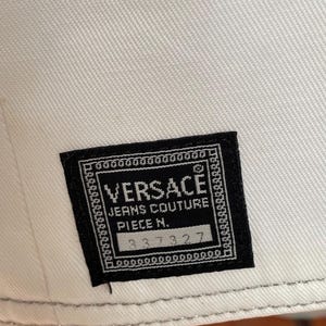 May include: Close-up of a white fabric with a black Versace Jeans Couture label. The label features the brand name, product details, and a serial number. The fabric has a subtle texture.