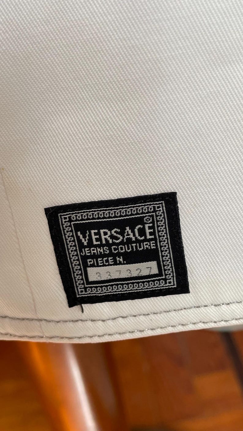 May include: Close-up of a white fabric with a black Versace Jeans Couture label. The label features the brand name, product details, and a serial number. The fabric has a subtle texture.