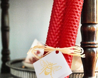 May include: Two red honeycomb beeswax candles tied together with a raffia ribbon and a white tag with a gold bee illustration. The tag reads "Honeycomb Candles".