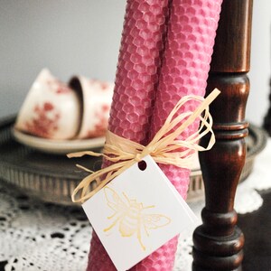 May include: Two pink beeswax candles with a honeycomb pattern, tied with raffia and a tag featuring a gold bee illustration. The candles are cylindrical and stand upright. The background includes a decorative table and vintage teacups.