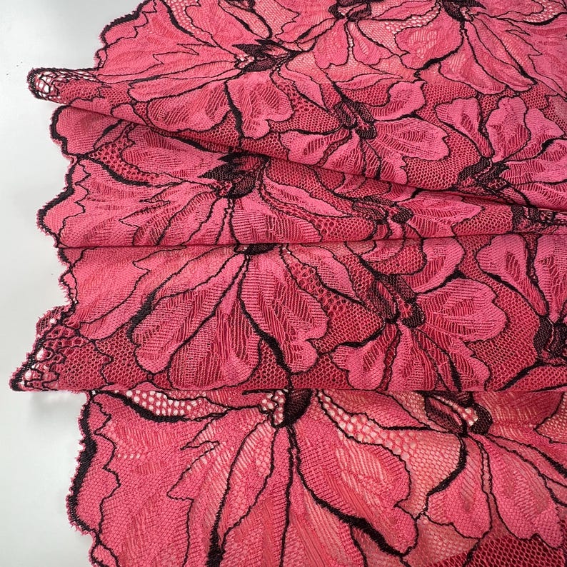Op de afbeelding: Close-up van gevouwen roze kanten stof met een bloemenpatroon. Het kant heeft zwarte contouren die contrasteren met het roze. De stof is gelaagd en toont de gedetailleerde textuur en het ontwerp.