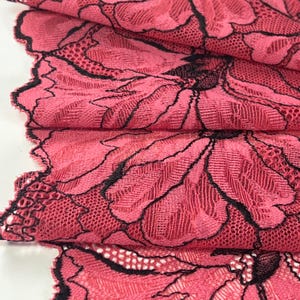 Op de afbeelding: Close-up van een gevouwen stuk roze kanten stof met een bloemenpatroon. Het kant heeft een gedetailleerd patroon met zwarte contouren, wat contrasteert met de roze achtergrond. De randen van de stof zijn geschulpt.