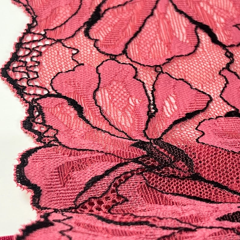 Op de afbeelding: Close-up van een gedeelte van roze kant met een bloemmotief. Het kant heeft een delicate, open weefsel met zwart dat de bloemvormen omlijnt. De randen zijn geschulpt, wat een decoratief tintje geeft. Het algehele effect is ingewikkeld en vrouwelijk.