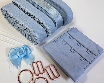 May include: A sewing kit containing light blue elastic bands, a matching hook-and-eye closure, two rose gold metal rings, two rose gold sliders, and a small light blue satin ribbon bow with a central rose detail for garment construction.