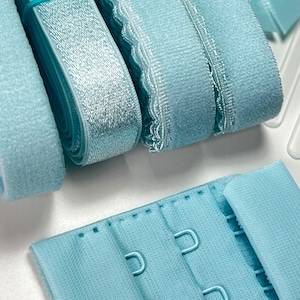 May include: Close-up of various light blue bra-making supplies. The image shows rolls of elastic, hook and eye closures, and other materials used in lingerie construction. The materials are arranged on a white surface.
