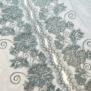 以下が含まれることがあります： 繊細な水色のレース生地のクローズアップ。精巧な花の刺繍が施されています。 大きく詳細な花と渦巻き模様のデザインで、スカラップエッジが付いています。 レースは透明感のある白い背景にあります。