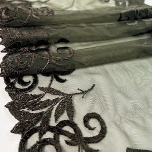 Peut inclure: Gros plan sur un tissu vert olive transparent avec une broderie florale marron foncé complexe. La dentelle délicate présente des motifs détaillés de feuilles et de volutes, créant un design texturé et élégant. Le tissu est plié, montrant des couches de matière transparente.