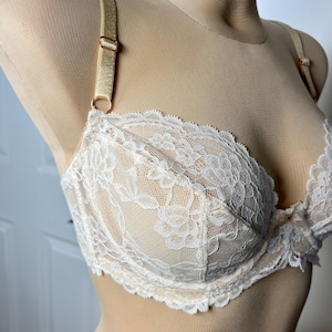 May include: A beige underwire bra featuring white floral lace overlay on the cups. The garment includes adjustable satin-finish shoulder straps with rose gold-toned metal hardware, a scalloped lace edge along the bottom, and a small center bow.